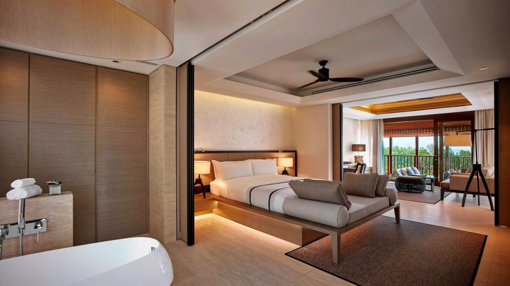 Modern hotel suite with a large bed, sofa, open bathtub, ceiling fan, desk, and sitting area near a balcony with trees visible outside through glass doors. Warm lighting and contemporary decor create a cozy ambiance.