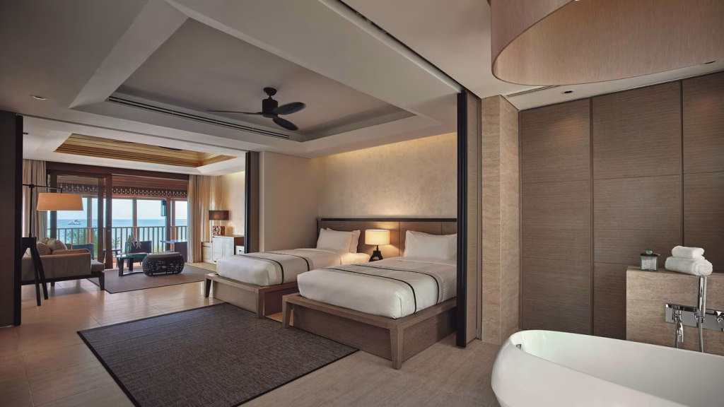 Spacious hotel room with two double beds, a sitting area with a sofa and lamp, a ceiling fan, large windows leading to a balcony with a view, and an open bathroom area with a bathtub and towels.