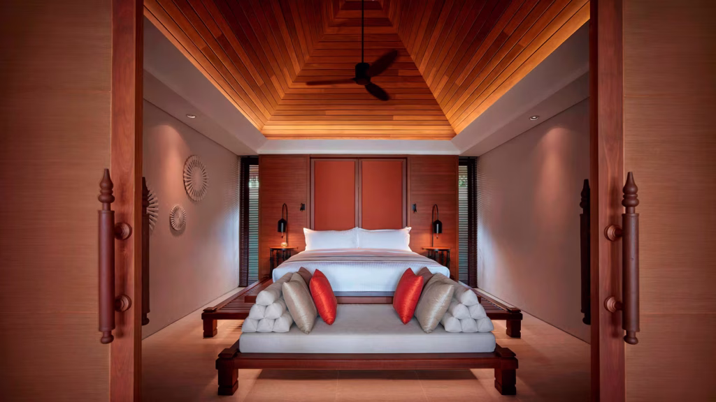 A luxurious bedroom with a high wooden ceiling, a large bed with white linens, a cushioned bench at the foot with red and gray pillows, warm lighting, and modern decor elements on the walls.