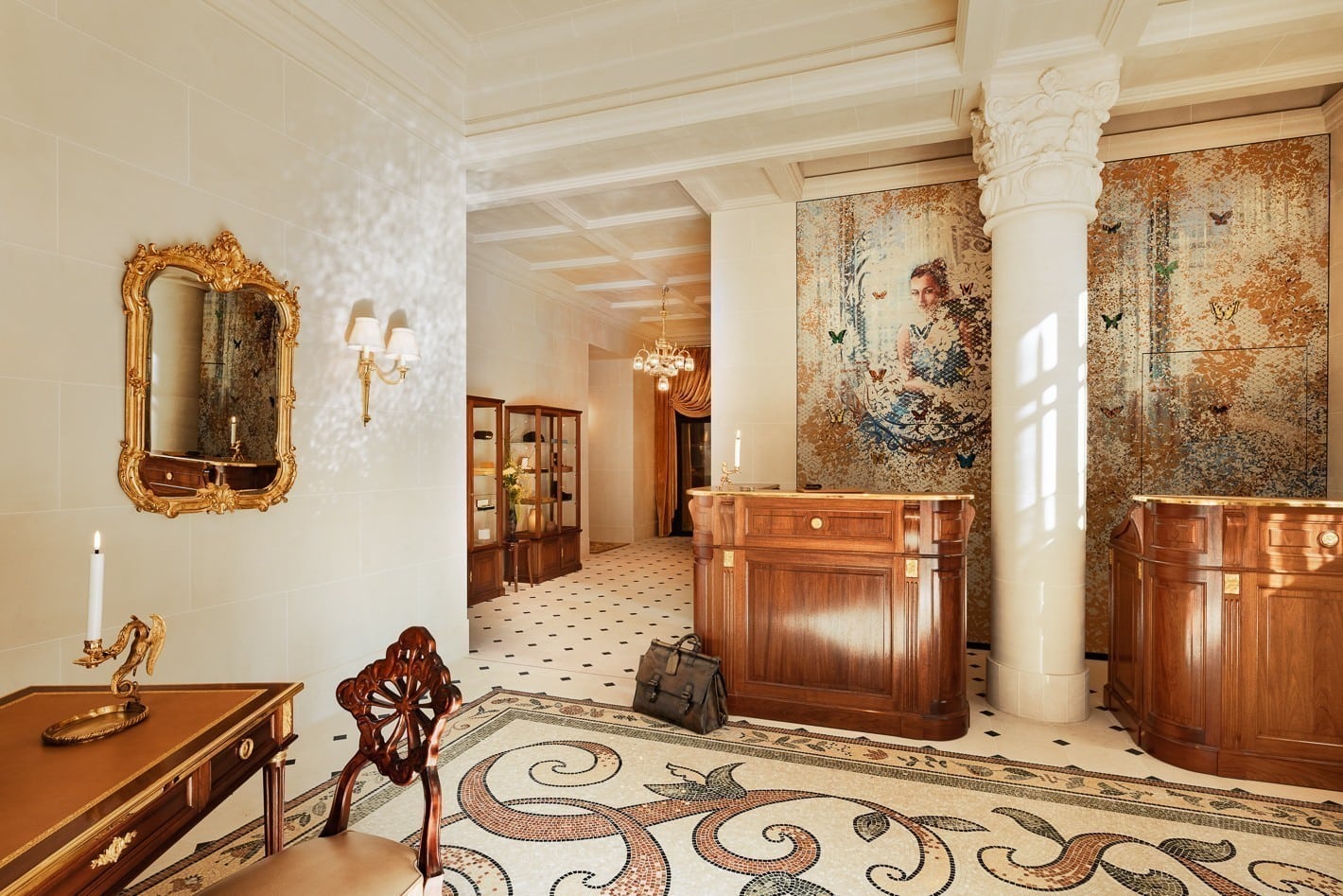 Elegant hotel lobby with ornate wooden desks, a mosaic floor, a gold-framed mirror, and a large mosaic artwork of a woman on the wall. A suitcase sits near the desk, and a lit candle is on a table in the foreground.
