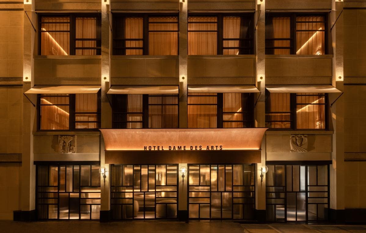 The warmly lit entrance of Hotel Dame des Arts at night, featuring large windows, illuminated awnings, art deco architecture, and soft amber lighting.