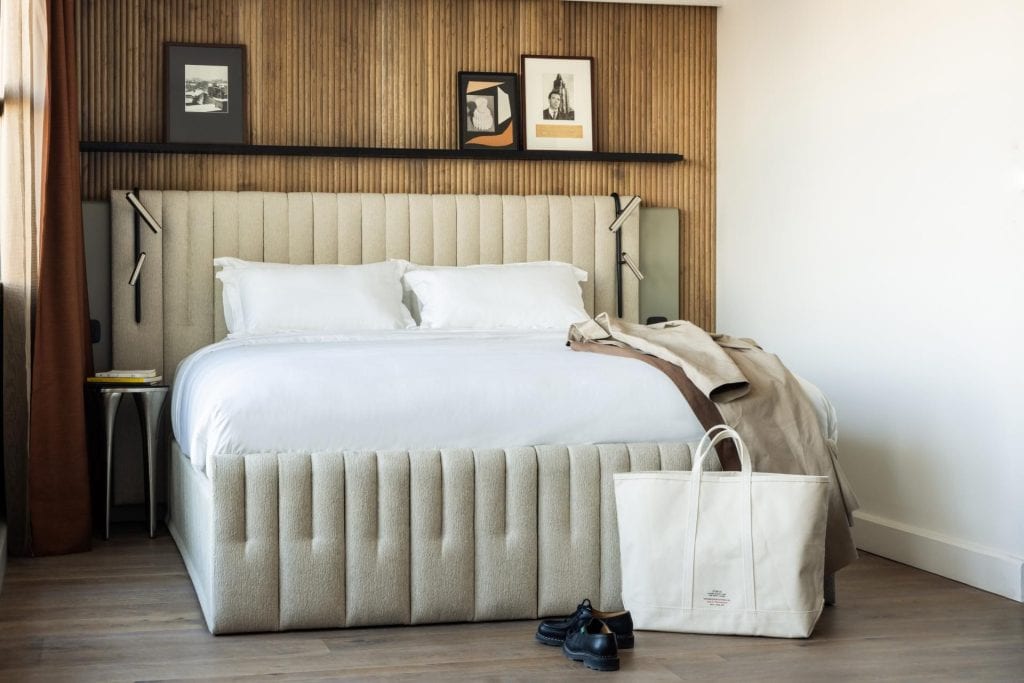 A modern, neatly made bed with a beige upholstered headboard is centered in the room. A beige coat, black shoes, and a large white tote bag are at the foot of the bed. Artwork hangs on a wooden accent wall behind.