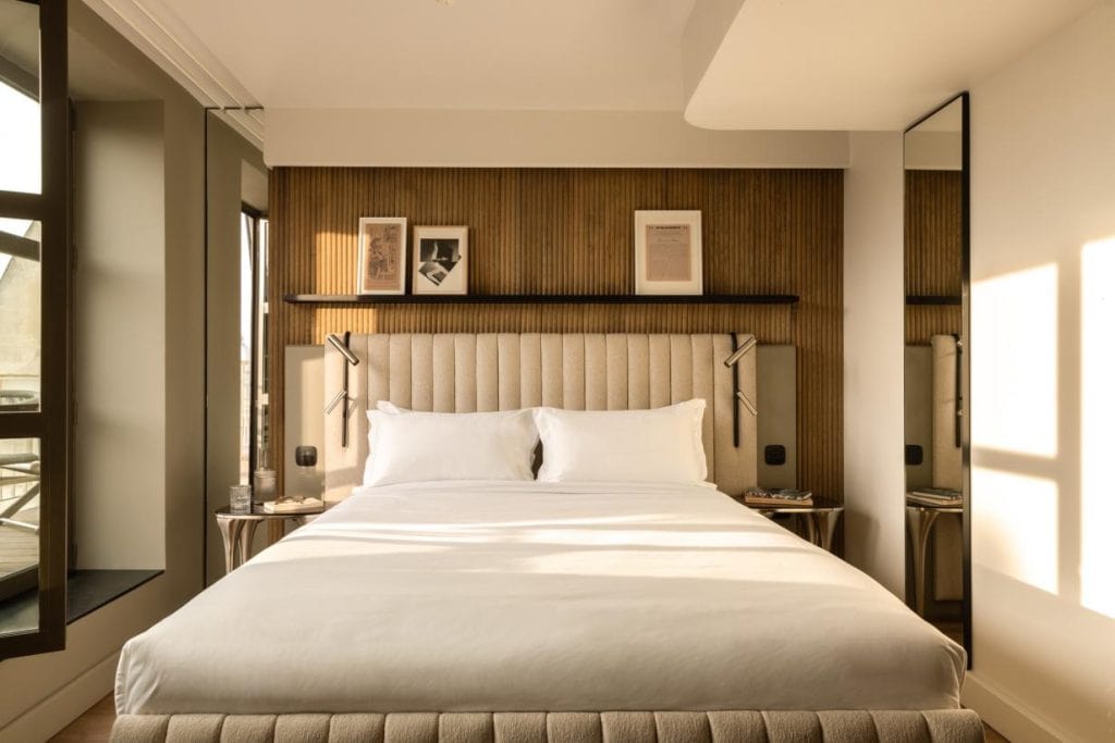 A modern bedroom with a large bed, beige upholstered headboard, and white bedding. There are framed pictures on a wooden accent wall, bedside tables with lamps, and large windows letting in natural light.