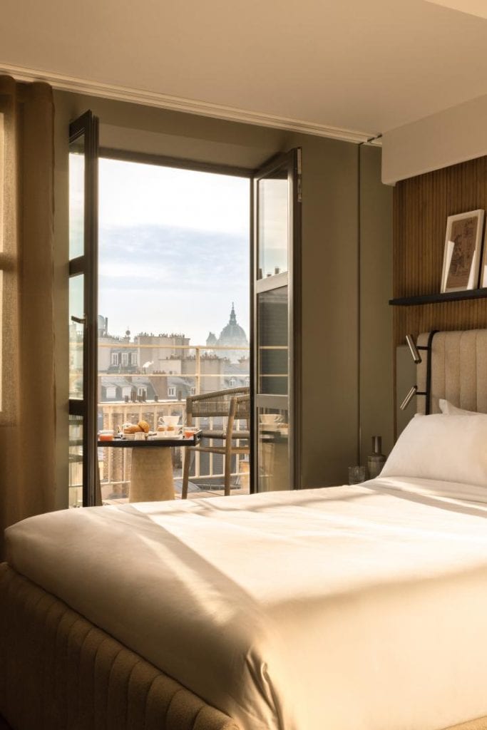 A cozy hotel room with a neatly made bed, sunlight streaming in through open glass doors leading to a balcony with a table and chairs, and city rooftops with a domed building visible in the distance.