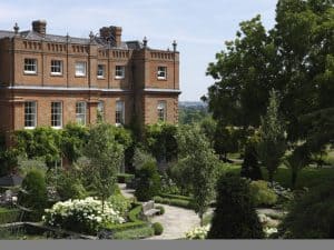 A large, historic brick building with tall windows stands behind a landscaped garden with stone paths, benches, flowering plants, and neatly trimmed greenery on a sunny day.