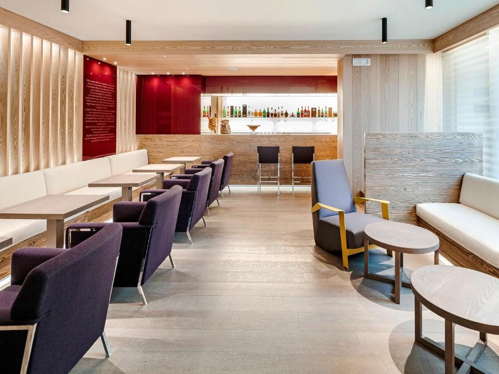 A modern lounge with light wooden floors and walls, purple and beige armchairs, small tables, and a sleek bar in the background with bottles displayed on red shelves. The space is well-lit and neatly arranged.