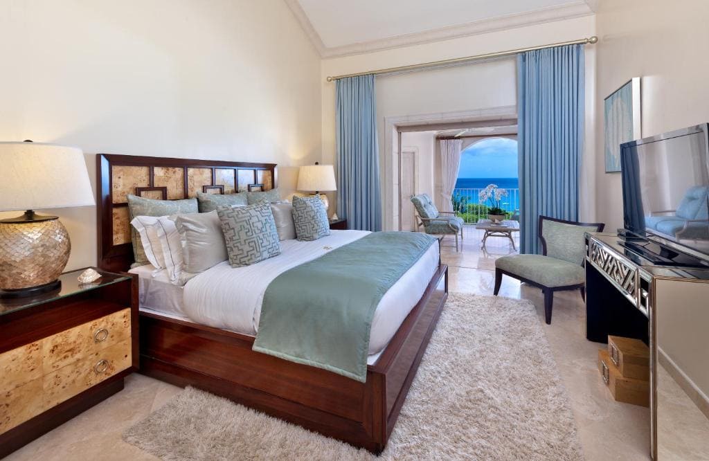 A modern hotel room with a large bed, pale blue and beige bedding, nightstands with lamps, a TV, and a balcony with ocean views through open curtains. The decor is light and airy with coastal accents.