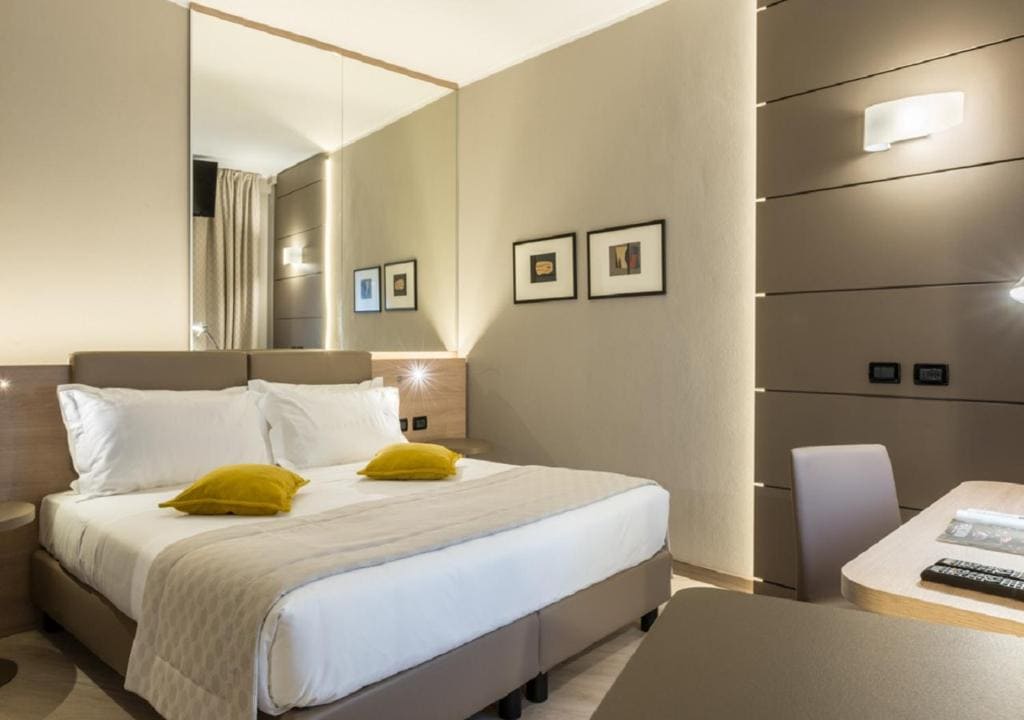 A modern hotel room with a double bed, white bedding, two yellow accent pillows, a mirror behind the bed, soft lighting, wall art, a desk, and neutral-toned walls and furniture.