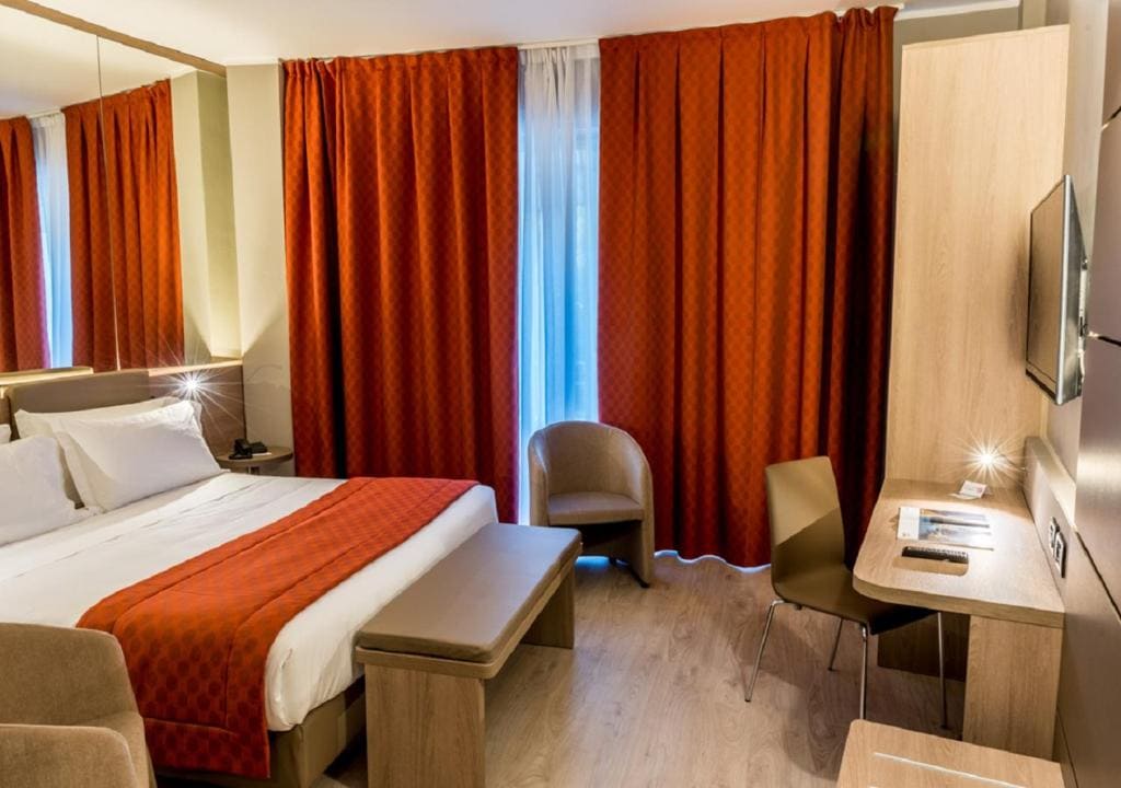 A modern hotel room with a double bed, red bed runner, matching red curtains, a bench, two taupe chairs, a desk with a chair, a wall-mounted TV, and light wood flooring.