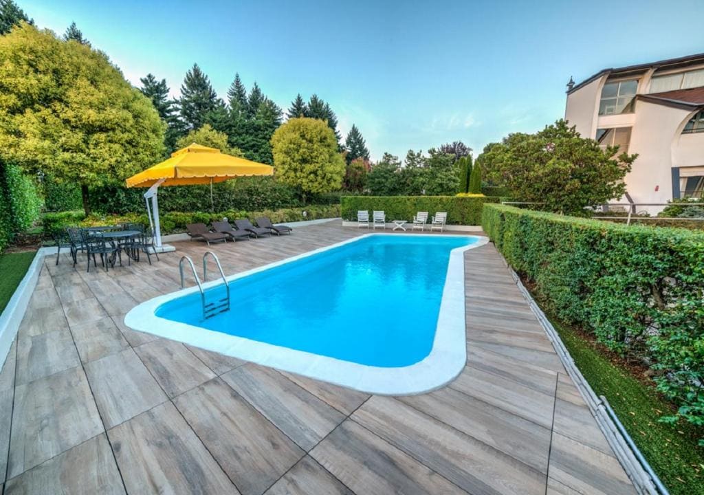 Rectangular outdoor swimming pool surrounded by a tiled deck, lounge chairs, a large yellow umbrella, table and chairs, and green hedges. Trees and a modern building are visible in the background.