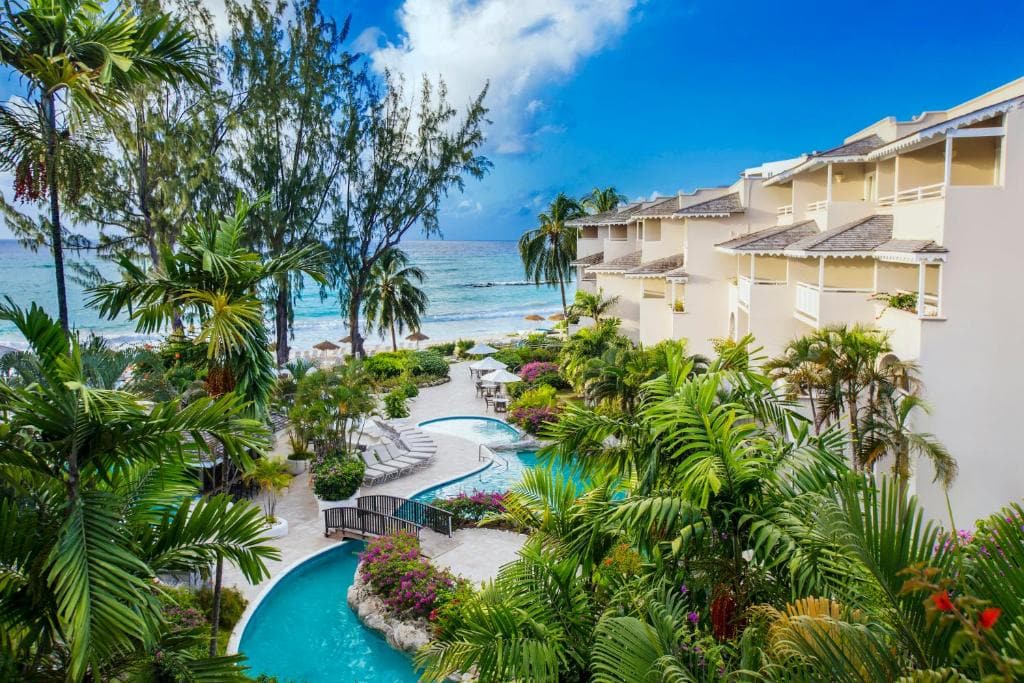 A tropical resort with cream-colored buildings, lush green palm trees, and a winding swimming pool, overlooks a sandy beach and the ocean under a bright blue sky with scattered clouds.