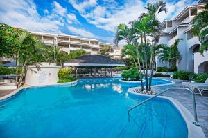 A tropical resort pool with clear blue water is surrounded by palm trees and white buildings under a sunny sky. A thatched-roof swim-up bar sits in the center, and lounge chairs line the pool’s edge.
