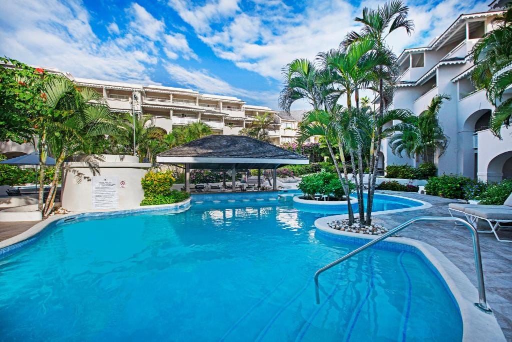 A tropical resort pool with clear blue water is surrounded by palm trees and white buildings under a sunny sky. A thatched-roof swim-up bar sits in the center, and lounge chairs line the pool’s edge.