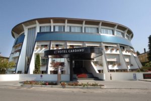 A modern, circular hotel building with blue and white exterior panels, Hotel Cardano. The entrance has steps, potted plants, a large sign, and windows adorned with flower boxes. The sky is clear and sunny.