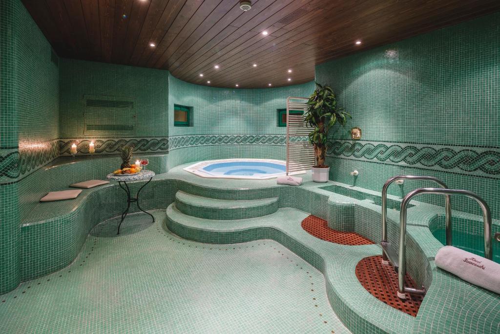 Luxurious indoor spa room with green mosaic tiles, a round jacuzzi, curved seating, candles, drinks on a table, a potted plant, and a towel warmer under a wooden ceiling with recessed lights.