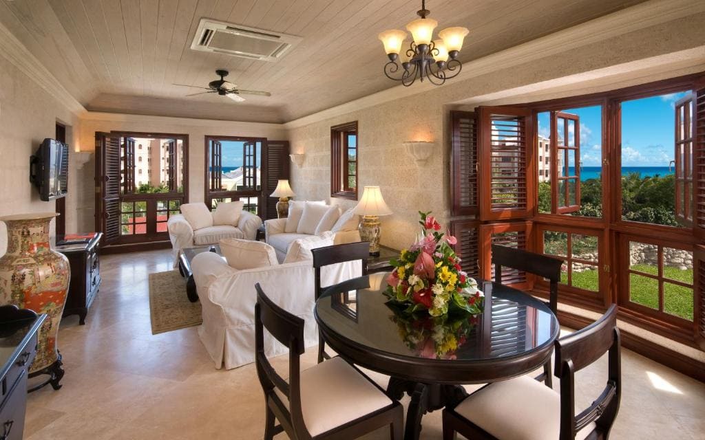 Bright, elegant living and dining area with white sofas, wooden furniture, and large windows with shutters, overlooking a green garden and ocean view. A floral arrangement sits on the dining table.