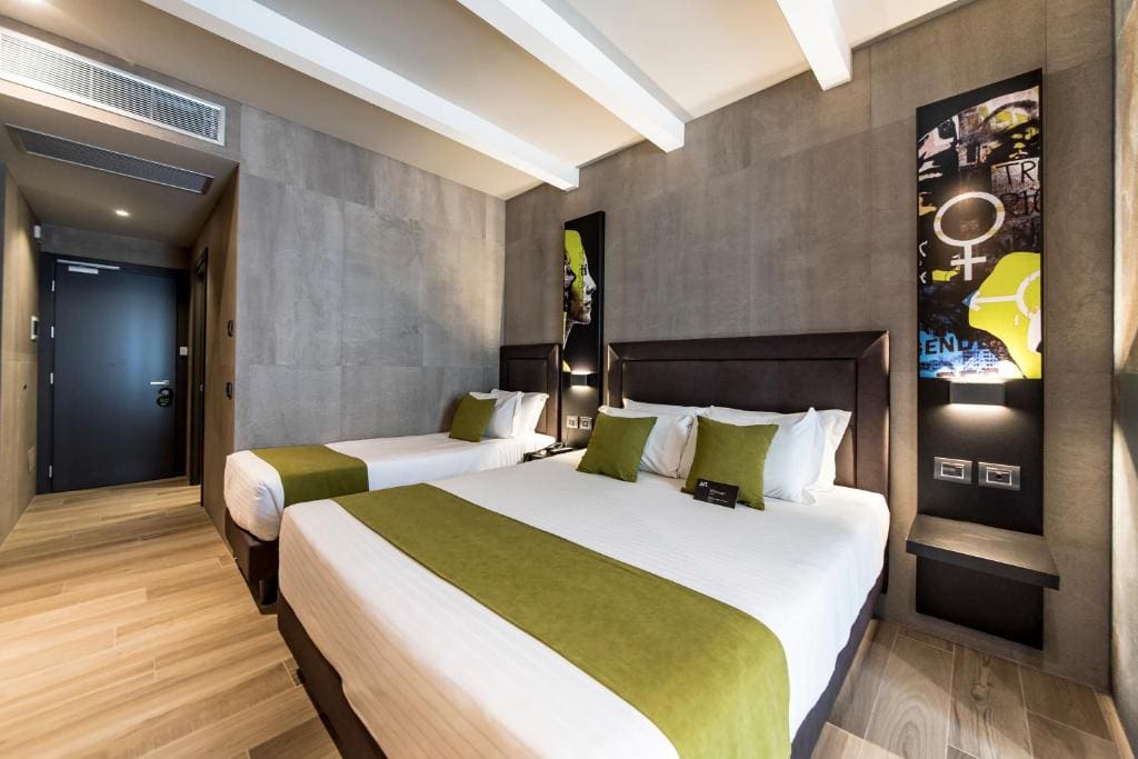 Modern hotel room with two beds featuring white linens and green runners and pillows, wood flooring, gray textured walls, colorful wall art above each bed, and a dark door at the end of the room.