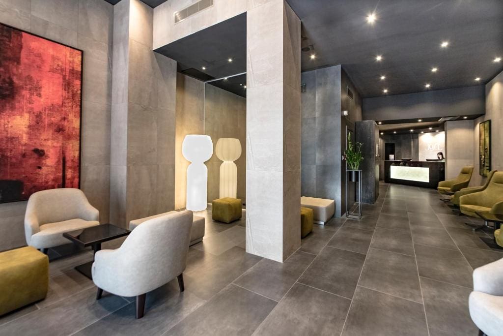 Modern hotel lobby with gray tiled floors and walls, beige armchairs, olive green ottomans, a large red abstract painting, unique white floor lamps, and a reception desk in the background.