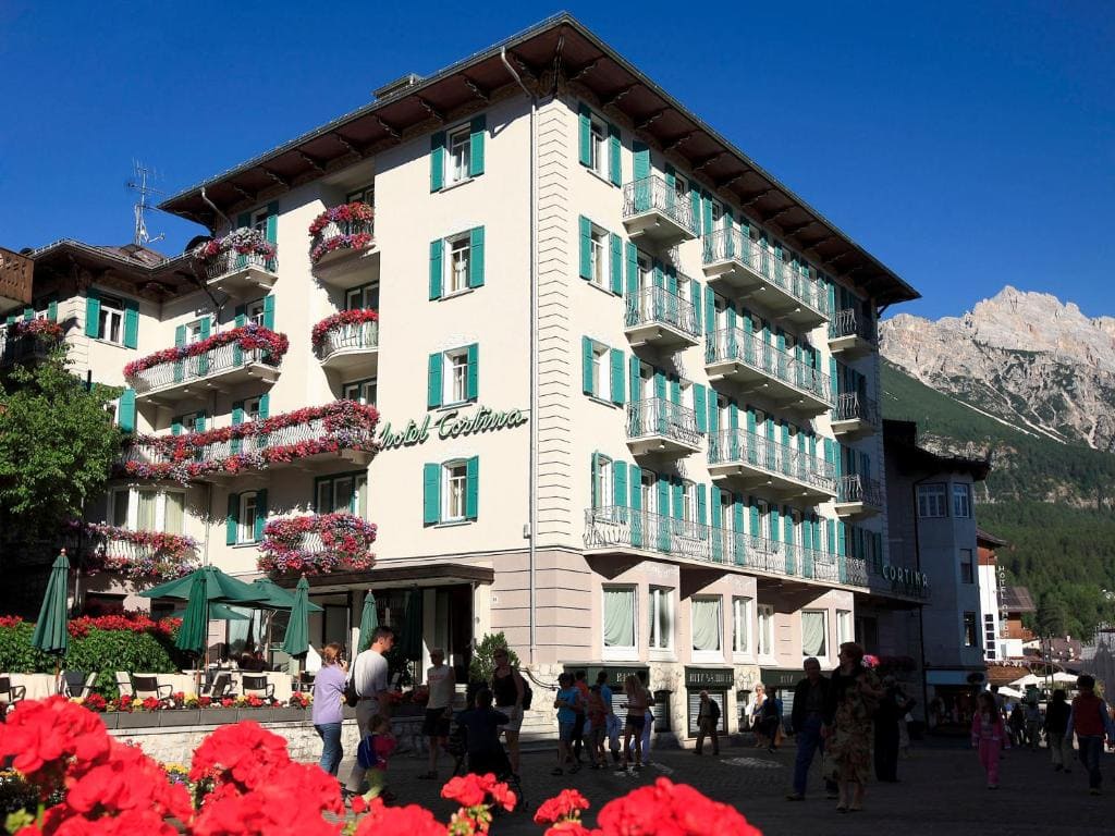 A large hotel with teal shutters and balconies adorned with red flowers stands on a sunny street, with people walking and sitting at an outdoor café; mountains are visible in the background under a clear blue sky.