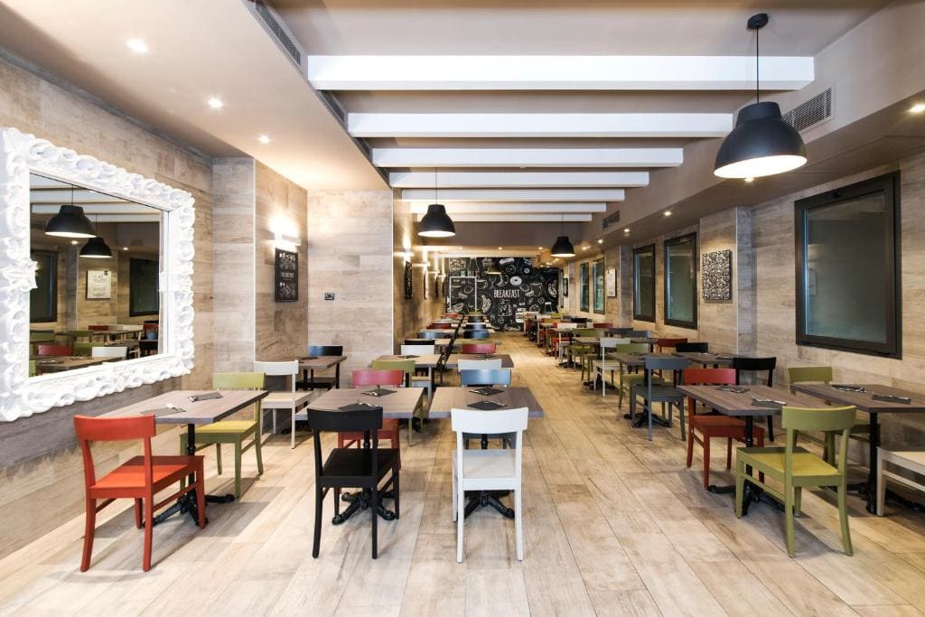 A modern, brightly lit restaurant with wooden floors, colorful chairs, black tables, large windows, a framed mirror on one wall, and chalkboard art at the far end. The space is empty and neatly arranged.