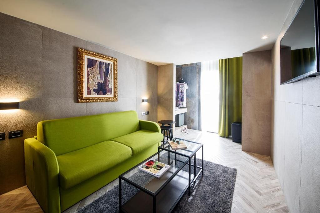 Modern living room with a bright green sofa, abstract artwork on the wall, a glass coffee table with magazines, gray rug, wall-mounted TV, and a window with green curtains letting in natural light.
