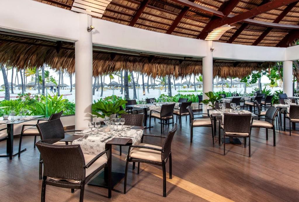 Open-air restaurant with wicker chairs and tables set with white linens, surrounded by lush green plants. Large windows overlook palm trees, a sandy beach, and thatched-roof structures by the water.