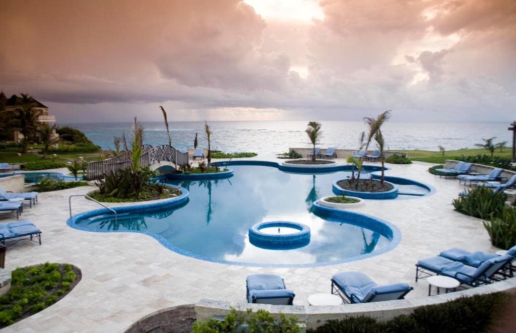 A luxurious resort pool with curved shapes, surrounded by blue lounge chairs and palm trees, overlooks the ocean at sunset under a dramatic cloudy sky.