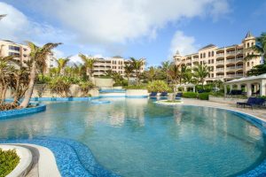 A large, curving outdoor swimming pool surrounded by palm trees, tropical plants, lounge chairs, and multi-story resort buildings under a partly cloudy sky.