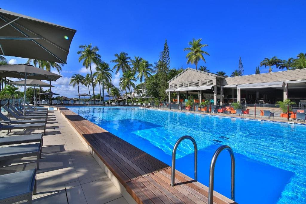 A large outdoor swimming pool surrounded by lounge chairs and umbrellas, with palm trees and a resort building in the background under a bright blue sky.