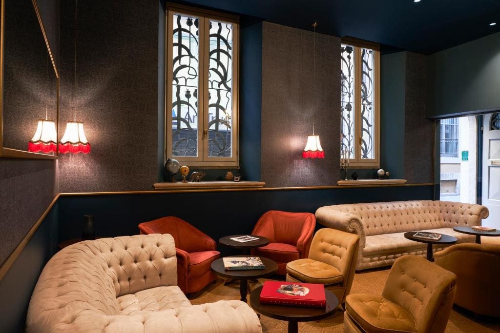 Cozy lounge with vintage armchairs and sofas, round tables with books, two tall windows with decorative grilles, and pendant lamps with red shades, all in a warmly lit room with dark blue walls.