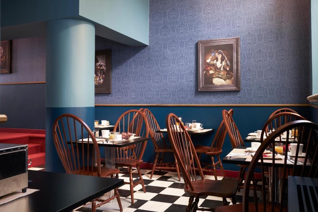 A small dining area with wooden chairs and tables set for breakfast, including cups, plates, and food. The room has blue walls, checkered floor tiles, and framed artwork hanging on the walls.