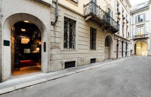 A narrow, quiet street lined with historic beige buildings featuring arched doorways, ornate ironwork, balconies, and tall windows; a warmly lit interior is visible through an open door on the left.