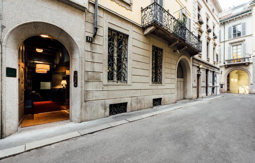 A narrow, quiet street lined with historic beige buildings featuring arched doorways, ornate ironwork, balconies, and tall windows; a warmly lit interior is visible through an open door on the left.