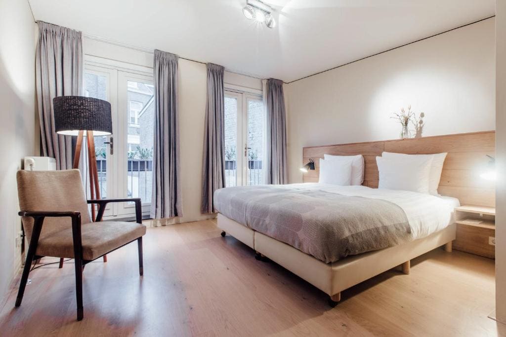 A modern, minimalist bedroom with a double bed, light wood floors, neutral bedding, a chair, a floor lamp, and large windows with gray curtains letting in natural light.