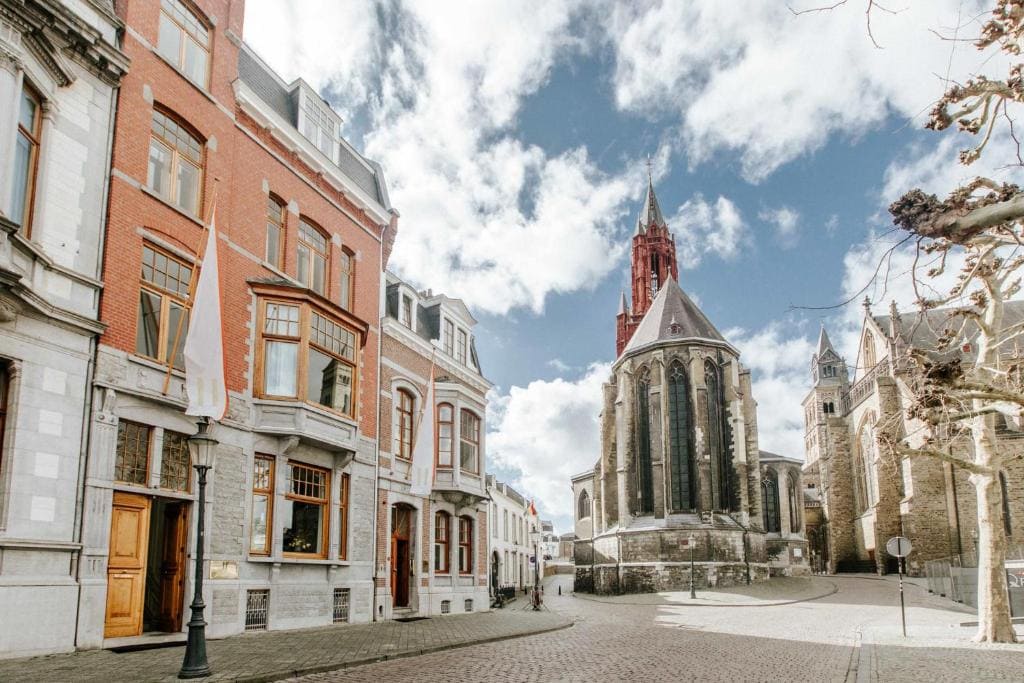 A cobblestone street lined with historic buildings leads to a large stone church with a tall, red steeple under a partly cloudy sky. Leafless trees and old architecture create a charming, European atmosphere.