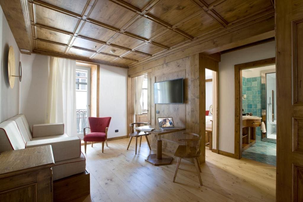 A cozy living room with wood-paneled ceiling and walls, a sofa, two chairs, a TV, and a small table. Natural light enters through a window. A bathroom with turquoise tiles is visible through an open doorway.
