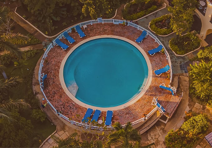 Aerial view of a circular swimming pool surrounded by brick-tiled decking and blue lounge chairs, with white fencing and lush green gardens around the pool area.