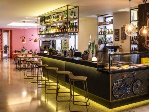 Modern café interior with a long bar counter, high stools, hanging glass racks, shelves with bottles, pendant lighting, and a display case with pastries. Dining tables and chairs are in the background. The atmosphere is warm and inviting.