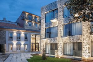 A modern building with an intricate, white lattice façade stands next to a renovated traditional stone house with balcony and warm lighting. Both buildings are illuminated at dusk, and a landscaped path runs between them.