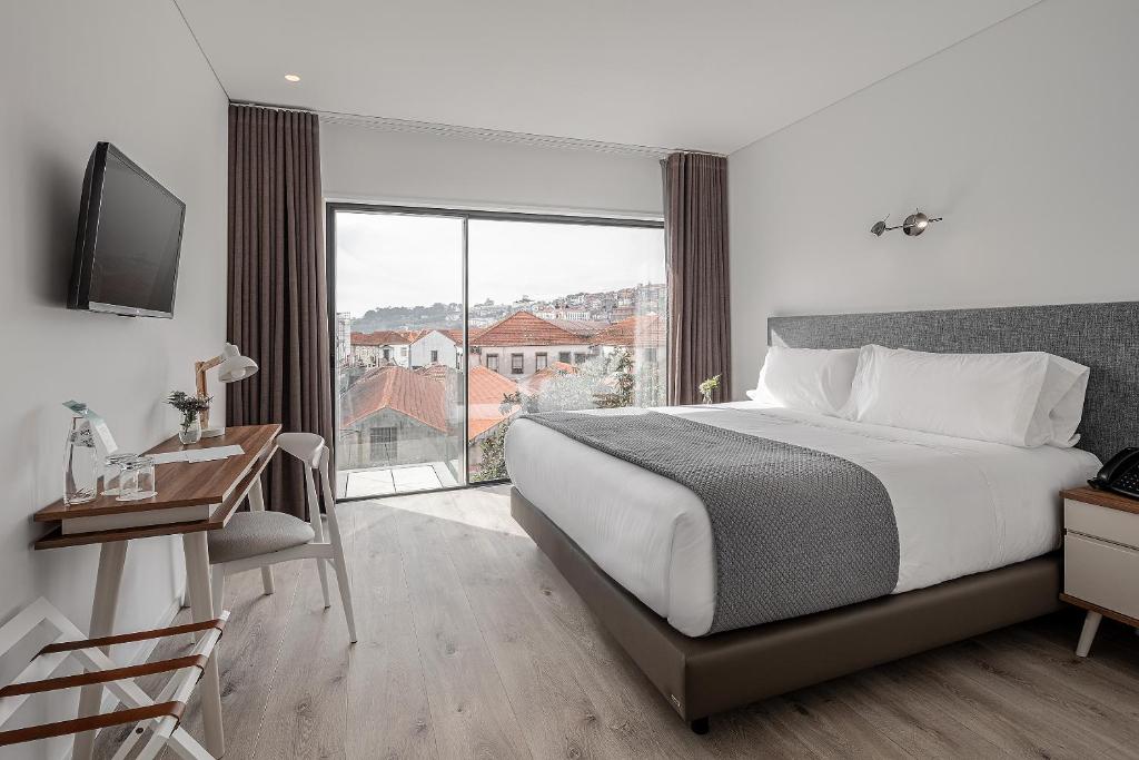 A modern hotel room with a large bed, desk, wall-mounted TV, and a glass door leading to a balcony with a view of red-roofed buildings and hills outside. The room has neutral colors and contemporary decor.