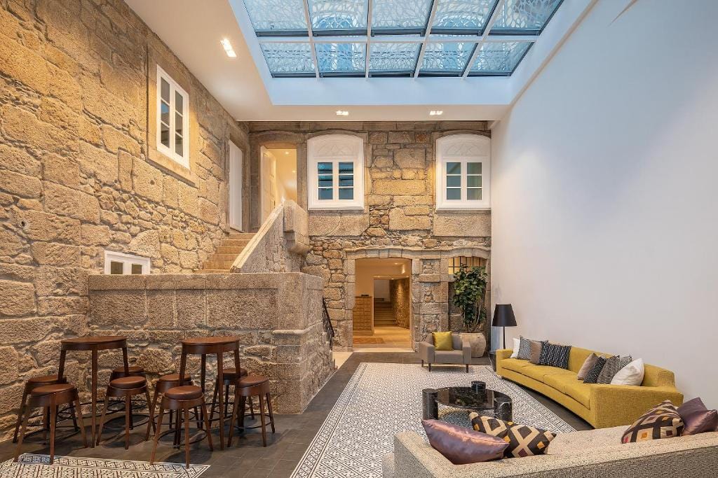 A spacious living area with stone walls, tiled floors, a yellow sofa, armchairs, bar stools, and a glass ceiling skylight letting in natural light. A stone staircase leads to windows and a hallway.