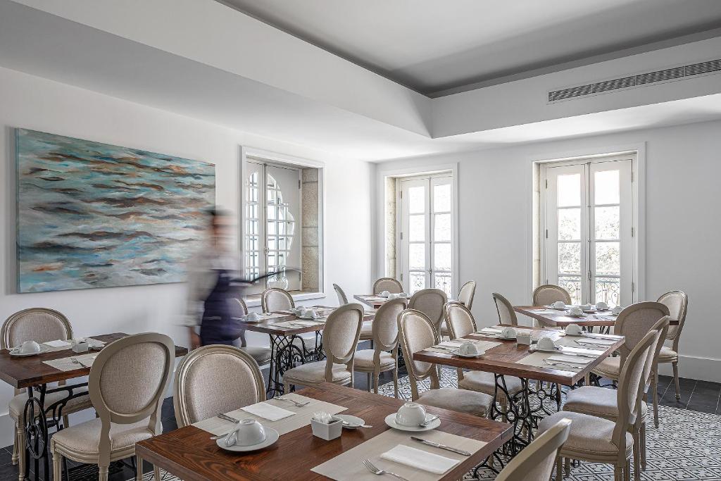 A bright, elegant dining room with wooden tables, beige chairs, white place settings, large windows, a blurred figure walking, and an abstract painting on the wall.