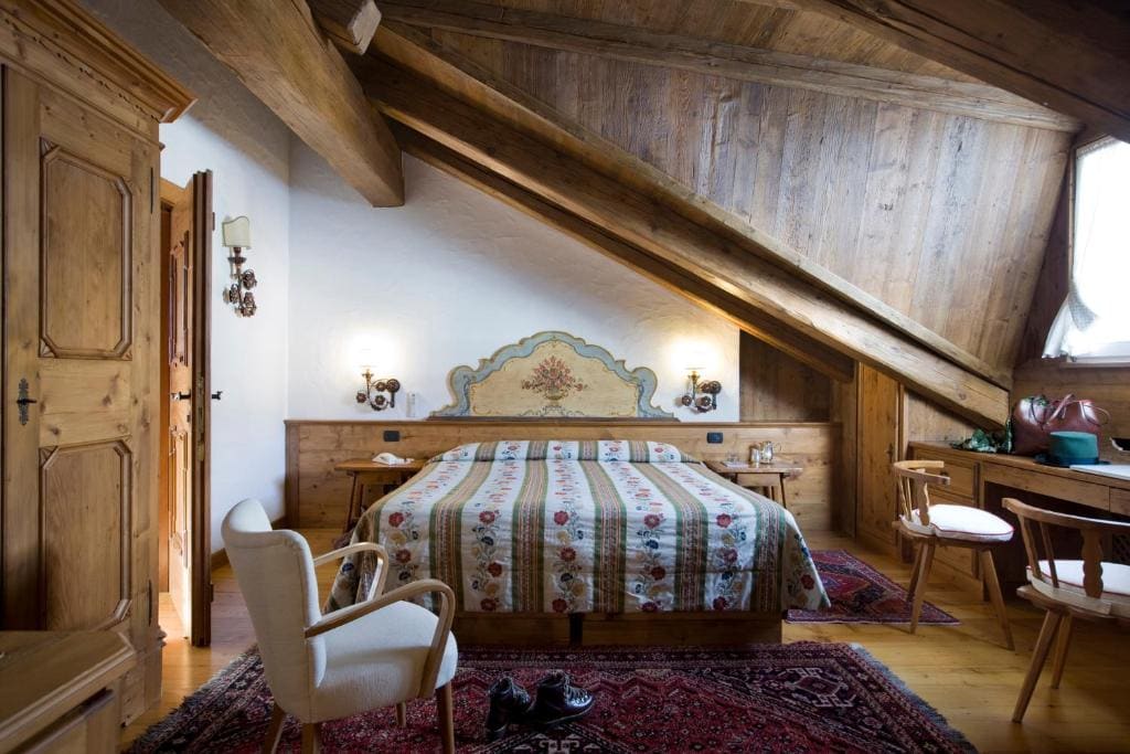 Cozy attic bedroom with slanted wooden ceiling, a double bed with a floral quilt, vintage wooden furniture, a small armchair, desk, wall sconces, and a patterned rug on wooden floor. Warm, rustic atmosphere.