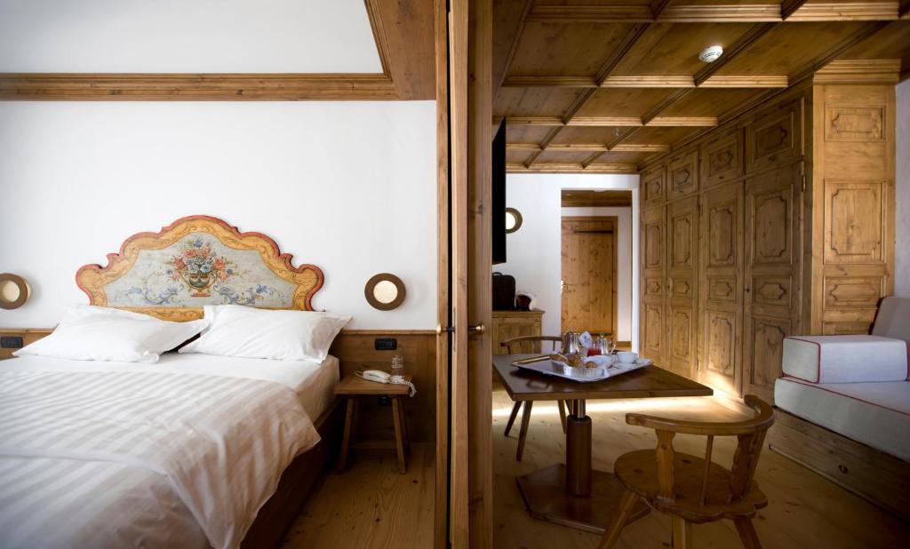 A cozy hotel room with a double bed featuring a decorative headboard, wooden furniture, and paneling. There is a small round table with chairs, a sofa, and neatly arranged refreshments on the table.