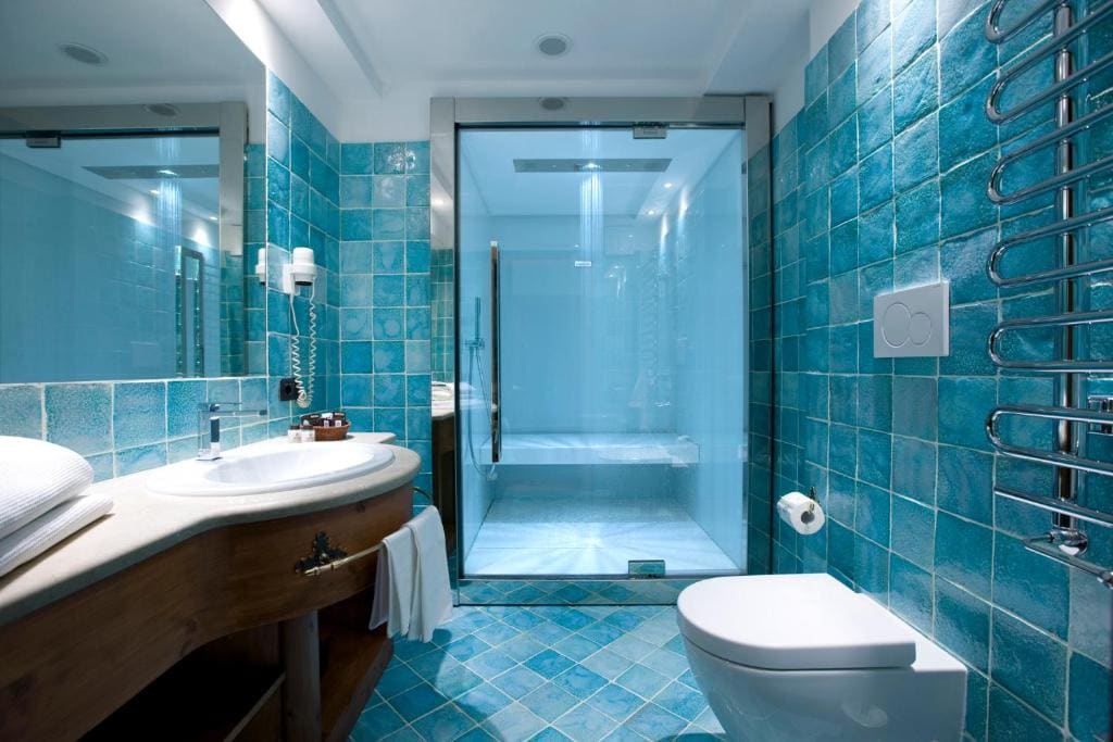 Modern bathroom with turquoise blue tiles, glass-enclosed shower, wall-mounted toilet, wooden vanity with white sink, large mirror, towel rack, and bright lighting. Clean and spacious design.