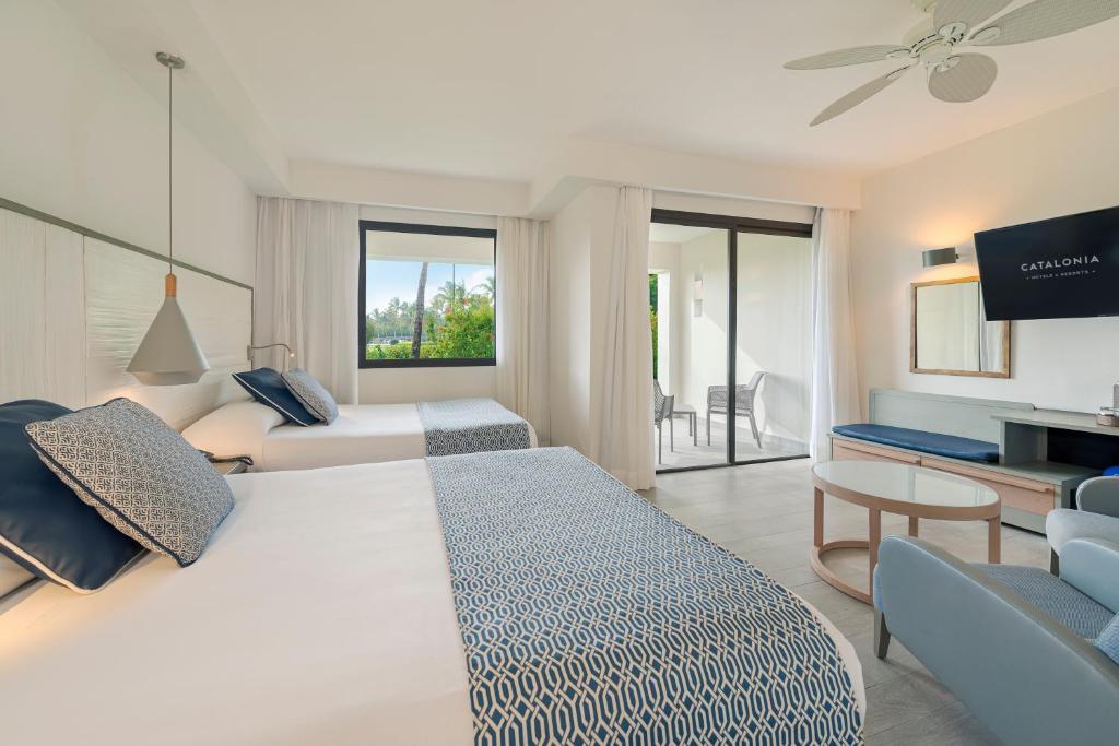 A bright hotel room with two double beds, blue and white bedding, a TV, a small sofa, a round coffee table, a ceiling fan, a desk, and a sliding glass door leading to a balcony with outdoor seating.