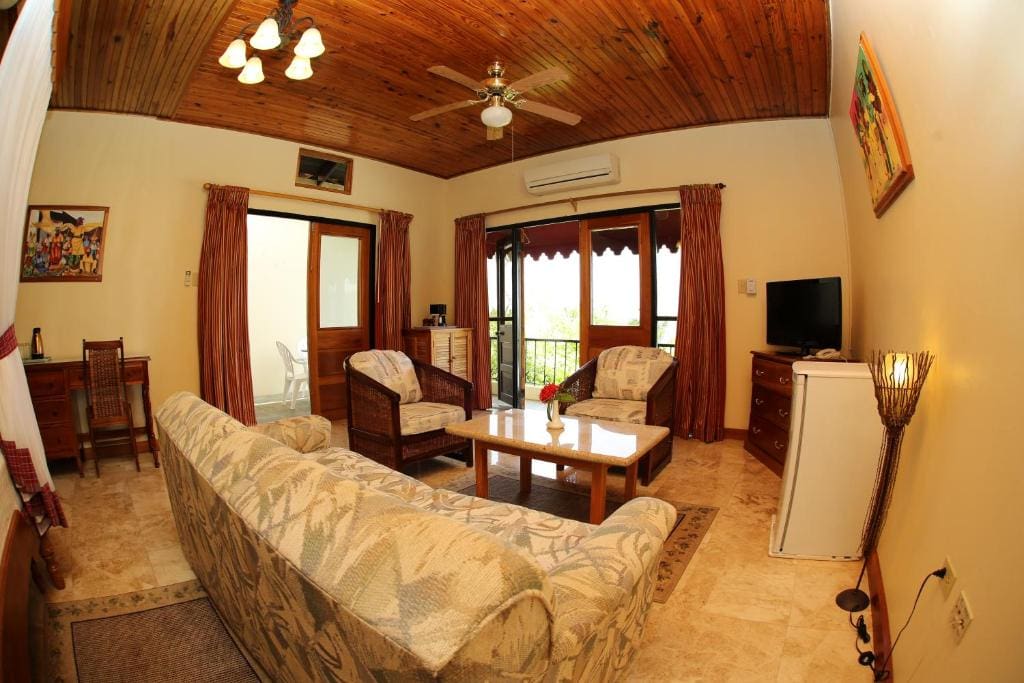 Cozy living room with wooden ceiling, ceiling fan, beige sofa, two armchairs, coffee table, TV on a chest, mini fridge, desk, and large windows with curtains letting in natural light.