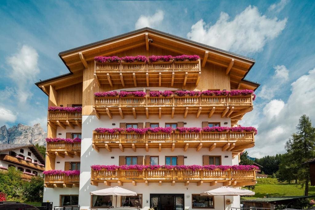 A wooden alpine-style building with four balconies, each decorated with vibrant pink flowers, set against a partly cloudy sky and green surroundings.