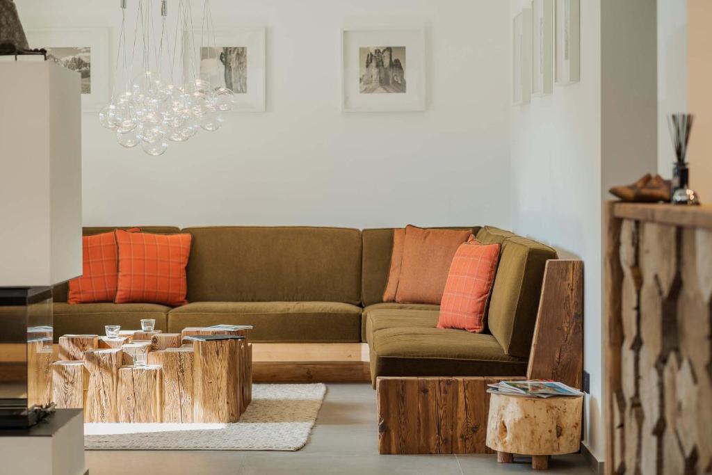 A modern living room with an olive-green sectional sofa, orange cushions, rustic wooden coffee tables, glass pendant lights, framed wall art, and a decorative wooden accent piece on the right.