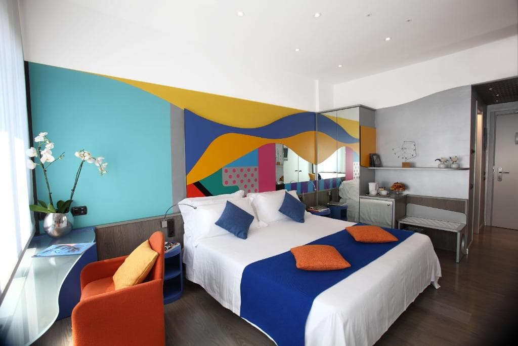 A modern hotel room with a colorful abstract mural behind a double bed, decorated with blue and orange pillows and throw. The room includes a glass desk, orange chair, orchid plant, and a large mirror.